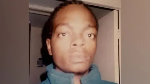 GMP Julius Matera, with black braided hair and wearing a blue-green jacket, stares at the camera while standing in front of an open white door