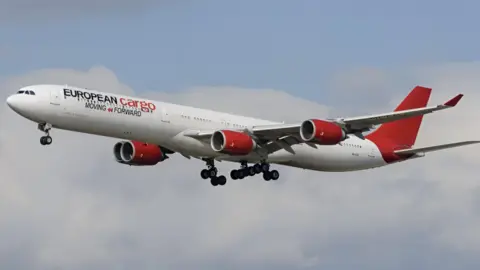 A white and red European Cargo plane in flight. Its wheels are down and there are clouds in the background. The body of the aircraft - which as a converted passenger plane still has windows along the side - is white. The tail is a completely red and, underneath the company name is the legend "moving forward". The aircraft has four jet engines, two on each wing, all painted the same red colour.