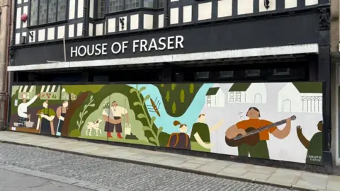 Charlie Stubbs / BBC It shows a shopfront, boarded up with colourful artwork of people cooking, collecting a bouquet of flowers, rowing in the river, and listening to a man busking. Above it is big white letters, with 'House of Fraser' in all capitals. It has an empty pavement in front of it with a grey brick road