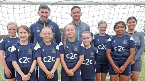 A team of young girls all lined up in front of a football goal and smiling. They are standing in front of Southend United boss Kevin Maher and captain Nathan Ralph. They are wearing blue kits.