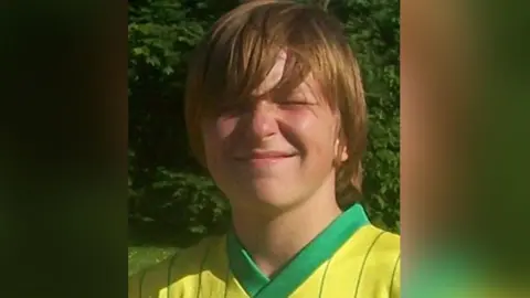 Beckie Ramsay The late teenager Dylan with long light brown hair wearing a yellow and green football shirt. He is smiling with his eyes shut as the sun is bright.