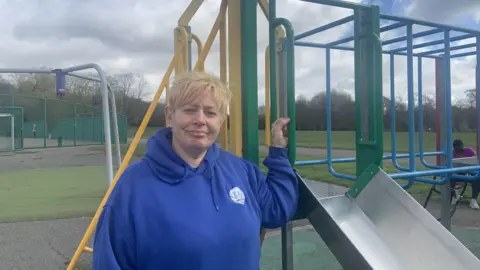A woman with short blond hair is wearing a bright blue hoodie and standing by a green and yellow climbing frame in a children's playground