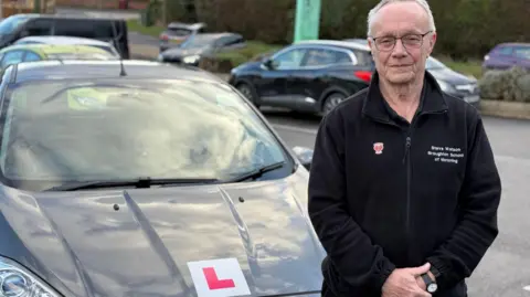 Driving instructor Steve Watson standing in a small car park next to a silver Ford Fiesta with a red and white L plate on the bonnet. Steve is wearing a black zip up fleece with 'Steve Watson Broughton School of Motoring' in white lettering printed on to the left side of his chest. He is in his sixties, wears black glasses and has short white hair. 