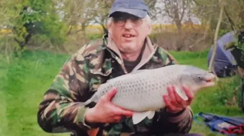 Family photo A man looks towards the camera while holding a large white and grey fish that he has caught. He is wearing a camouflage jacket and a blue cap. He is standing on a grassy bank with trees and a blue tent behind him.