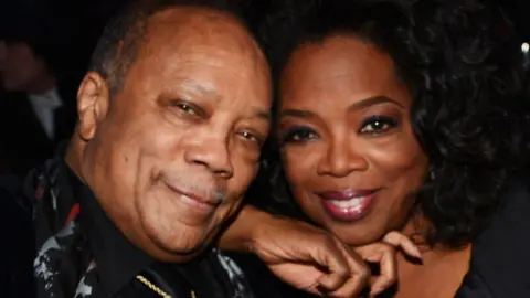 Quincy Jones and Oprah Winfrey sitting with their heads close together and smiling at the Rock and Roll Hall of Fame Induction Ceremony in 2013