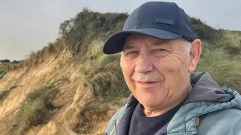 Andrew Turner/BBC Kenny Chaney stands near the eroded cliffs at Hemsby.