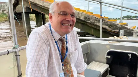 A man on board a ferry. He is wearing a white top with a red check pattern. His trousers are cream. He is smiling and wearing black glasses.