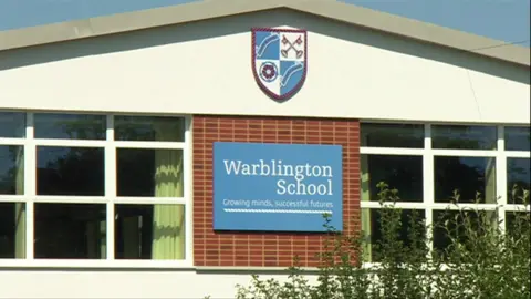 The front facade of Warblington School. Between two large windows is a large blue and white sign saying the school's name, with it's coat of arms logo above it