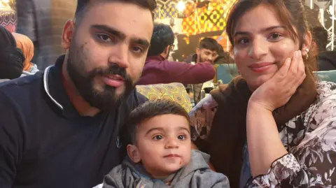 Hassan Naseer sits with his son next to his wife Sharzia Bibi, who is smiling with her head in her hand, which is propped up on a table. The family appear to be sat together at a restaurant. 