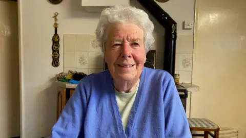 Pat Graham is looking at the camera lens. She wears a pale green t-shirt with a lavender blue cardigan on top. She is sitting in her farmhouse kitchen with a range behind her. On the wall are some horse brasses. 