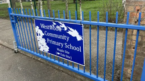 John Devine/BBC A blue gate has a sign on it which says Westwood Community Primary School in white writing on a blue background. There is a field in the background. 