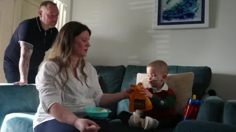 The image shows Jonah, who is three, sitting on a sofa at home and holding a paper bag with a doughnut in it. His mother is sitting next to him passing him the bag. His father is standing beside the sofa and looking at him. Jonah has a nasogastric tube fitted.