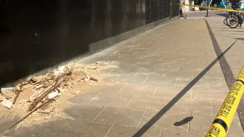 Owen Sennitt/BBC Debris can be seen on a street outside a department store on a hot summer's day, with yellow tape cordoning the area off
