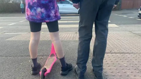 The legs of two school children, one walking and one on a scooter as they stand in front of a zebra crossing