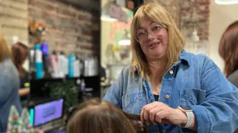 A woman with blonde hair wearing a denim jacket and doing someone's hair in a salon. She is looking and smiling into the camera wearing clear glasses.