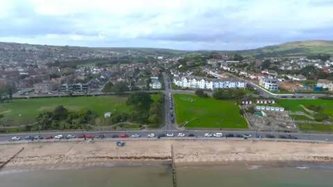 Aerial view of sea and sandy beach with cars parked along a seafront road and a large green area stretching behind.