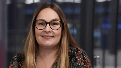 Sade Asker in the BBC Cymru Wales building with glass partition behind her. She is wearing a floral blouse and has glasses. Her chest length hair is brown with lighter highlights at the ends