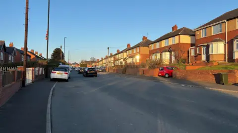 A grey tarmac road that is wide enough for two cars but with no paint markings on it. The road is bordered by red/yellow brick semi-detached houses with raised front gardens. There are cars parked on either side of the road. And about three cars driving into the distance.