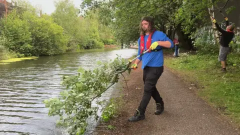 Man in a blue jumper and red life jacket pulling a piece of a branch out of the canal in Pleck, Walsall 
