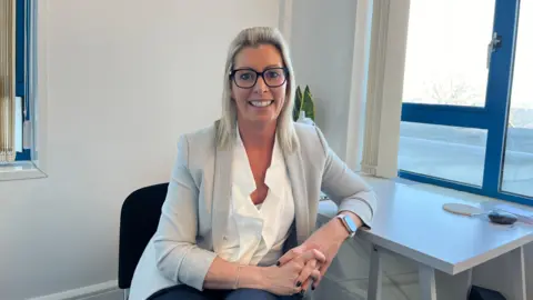 Dr Amy Gilmour, sitting at a desk and smiling at the camera