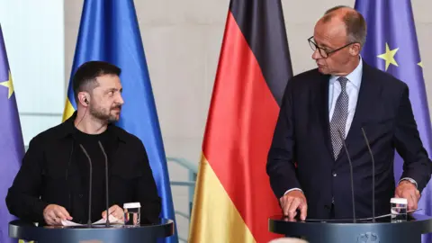 Volodymyr Zelensky and the German chancellor stand in front of two lecterns looking at each other. The flags of Ukraine and Germany are hanging behind them.