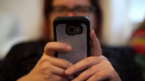 A woman holds out a mobile phone out towards the camera with both hands. Her face is blurred but she is wearing glasses and has dark hair.