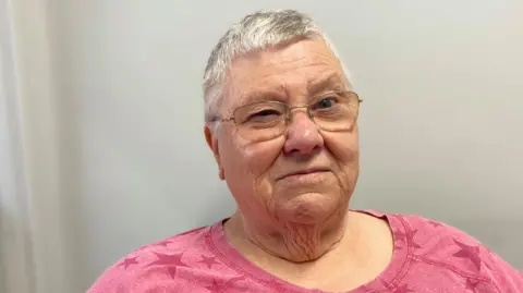 A woman with short grey hair and glasses is looking at the camera. She is wearing a pink top with a star pattern on it.