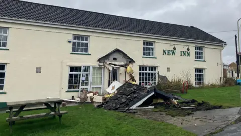 Front of New Inn pub with lettering attached to wall of long building painted cream colour. The front porch has been destroyed and debris is strewn across the path leading to the entrance.