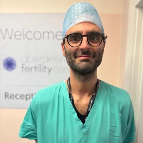 Dr Nicola Marconi wearing green scrubs, a black lanyard, dark glasses, and a light blue head covering. He's in front of an Aberdeen Fertility Clinic sign