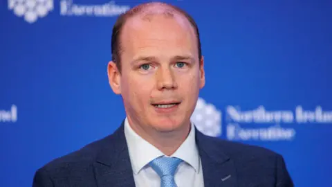 Gordon Lyons. He is mostly bald with a bit of short dark hair and blue eyes. He is wearing a dark blue blazer with white shirt and light blue tie. A blue background is behind him with NI Executive branding on it.