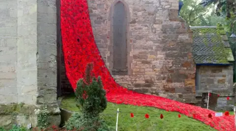 BBC Hundreds of red knitted poppies hang down the side of a grey stone and brick building with a narrow archway in the middle. Some poppies are also covering the grass.