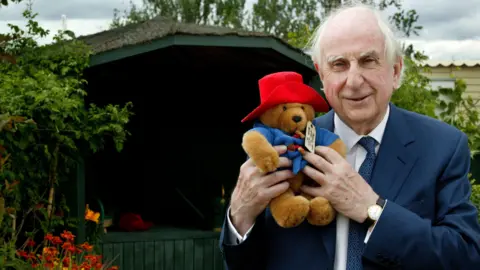 Author Michael Bond wearing a blue suit with a white shirt and blue tie. He is holding and a toy Paddington Bear who wearing a blue duffel coat and red hat. 