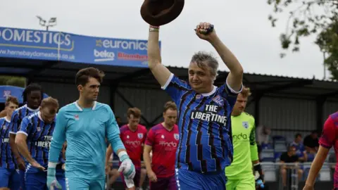 Pete Doherty raising his arms and a brown hat to the crowd as he leads out a football team in blue and black kits, which he is also wearing.