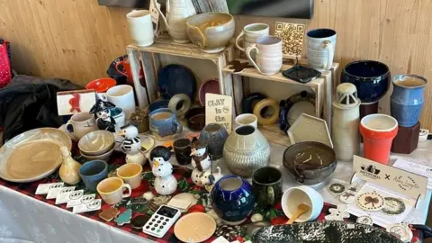 Alan Dunnighan A selection of different pieces of pottery including plates, cups and mugs in a variety of colours and styles. The items are arranged on a table and on wooden crates placed against a wooden wall.