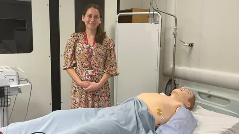 Courtney Masterson is standing in front of a hospital bed with a dummy in front of her wearing blue scrubs. Courtney is wearing a flower patterned dress and has brown hair. Her hands are folded in front of her.