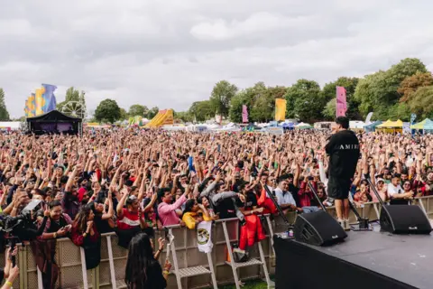 Sandwell & Birmingham Mela A man on stage sings into a microphone in front of hundreds of people in the audience.
