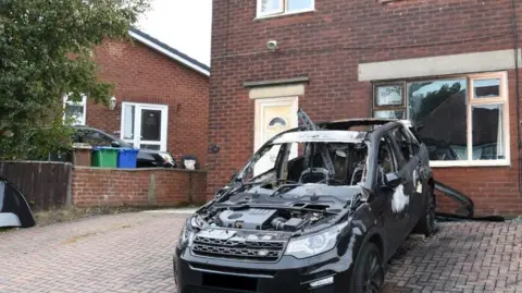 GMP Luanne Gill's black burnt out car in front of the fire damaged house with its cracked and smashed windows