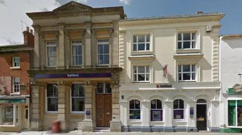 Google A two-storey stone building to the left with two large windows and a large wooden door on the ground floor. A white and cream building to the right with three storeys, all with large windows and white frames. There is a narrow dark-coloured door to the right.