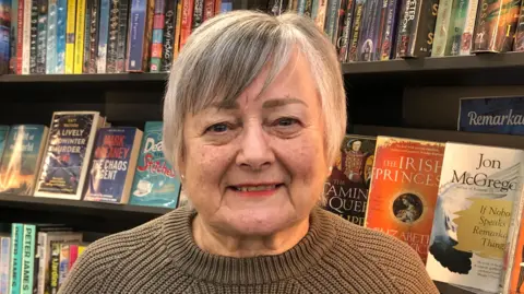 Sue Holden wearing a brown jumper.  She has short grey hair and is smiling at the camera.  She is standing in front of shelves of books at the community library she helps to run.