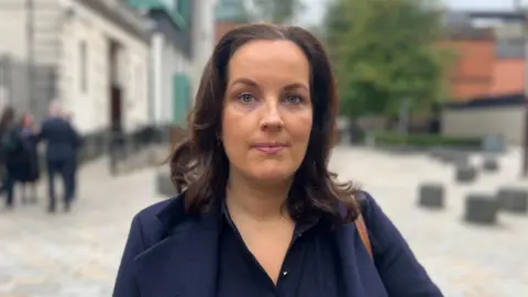 Claire McKeegan. She has shoulder-length dark brown hair and blue eyes. She is wearing a navy blazer with navy blouse.