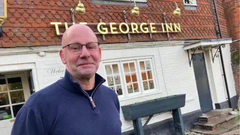 LDRS A bald man standing outside a pub. There is a gold sign on the pub which reads "THE GEORGE INN".
