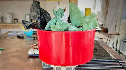 Gill Thomas Red bucket filled with black and green dog poo bags inside a kitchen. 