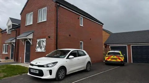Two police vehicles are parked on the drive of a newbuild house. One is a white car without any police markings on. A police officer is sat in the driver's seat. The other car has full police colours. There is a silver foil sheet over the downstairs window of the house.