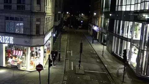 Met Police CCTV showing a wide shopping street in London at night. Three men are on the pavement, all wearing black.