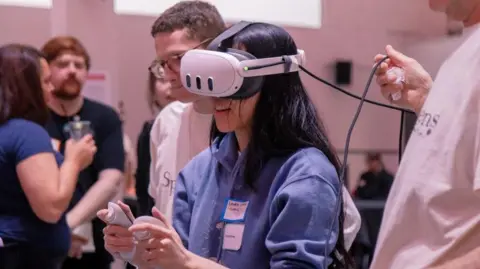 Corrupted Visuals A woman wears a virtual reality headset while holding a controller in each hand. Two people are standing by her and watching, while three people are blurred out as they talk to each other in the background.