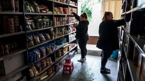 Coventry Foodbank Pathfinder support service at risk of closure 1 Getty Images Two workers in a food bank organise supplies on shelves, which have supplies of tinned and dry food. There is a step on the floor to reach the higher shelves and a window behind the workers.
