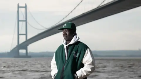 Anei smiles as he looks ahead, with the Humber Bridge in the background. Anei is wearing a bottle green cap and matching hooded jacket, blue jeans and white trainers.