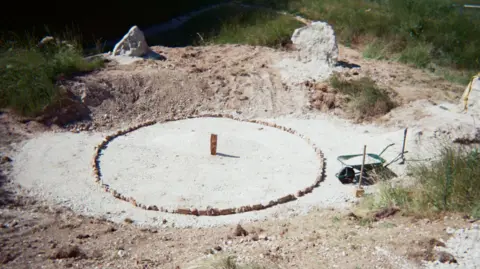 Matthew Rosier A circle of small stones with a rock in the middle. Outside the circle is a wheelbarrow full of chalk and there is an outer circle of big chalk stones.