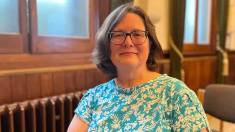 A woman with glasses and dark brown hair just above the shoulders. She is wearing a turquoise and white flowered t-shirt. She is in an old hall with wood panels, windows and a radiator behind her.