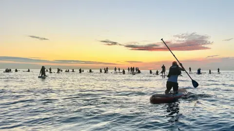 BBC A large number of paddleboarders on the water as the sun rises.